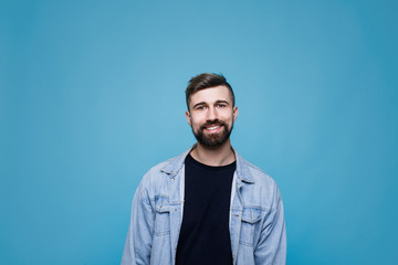 Obraz premium Young cheerful man in black t-shirt and denim jacket. Guy with beard expresses happiness. Blue background.