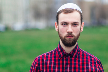 Portrait of religious young Jewish guy in traditional jewish male headdress, hat, boom, or yiddish on his head. Serious Israel man with beard looking at camera outdoors. Copy space, place for text.