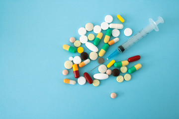 Multi-colored pills and capsules placed on a blue-green background with a syringe in the center of the conceptual image