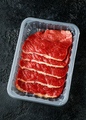 Raw fresh sizzling beef steak slices in plastic tray in butcher's shop on dark background table