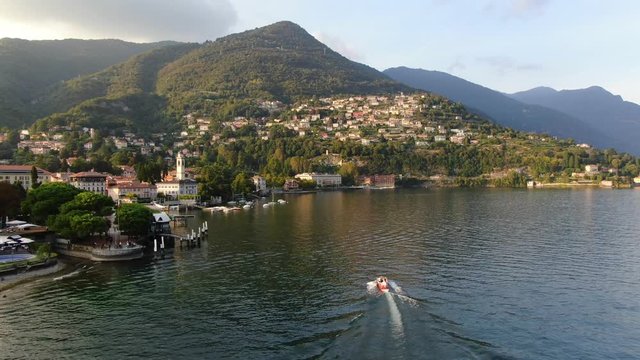 A red boat with the bride and groom is sailing towards the town of Cernobbio - the famous Italian resort. Luxury villas and hotels. Aerial view, shooting from a drone on a summer sunset day.