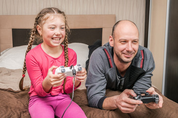 A father and a little daughter play video games using gamepads at home. Man and a child hold game joysticks in their hands. The family is having fun in the bedroom while being quarantined.