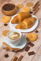 Breakfast with coffee cup, croissants, cookies, chocolate, coffee beans and cinnamon on wooden background. Latte art.
