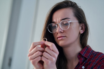 Frustrated pensive divorced woman with sad eyes holds ring and sits alone at home after break up relationship and divorce