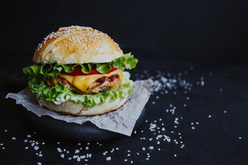 Appetizing fresh homemade burger, with chicken cutlet, lettuce, tomatoes, cheese and sauce. On a wooden board on a black background