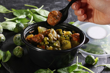 Healthy green food. Stewed cabbage, potatoes, peas and meatballs in a black plastic bowl with a spoon and hand. Green brussels sprouts, spinach on a black background. Background image, copy space