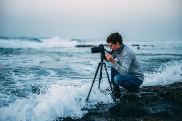 Obraz premium Young handsome photographer with curly hair, a gray sweater and jeans with a tripod and a camera on the beach, drenched in waves. Photography in Israel. Copy space. Shtorm on Mediterranean Sea