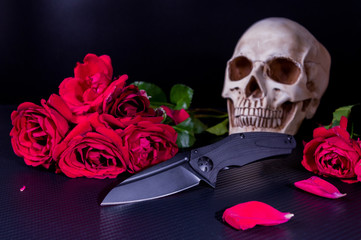Black folding knife on a background of red roses and a skull. Low key.