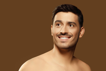 Handsome young man with naked shoulders looking aside with happy dreamful smile, concept of men beauty and skin care, isolated on brown background
