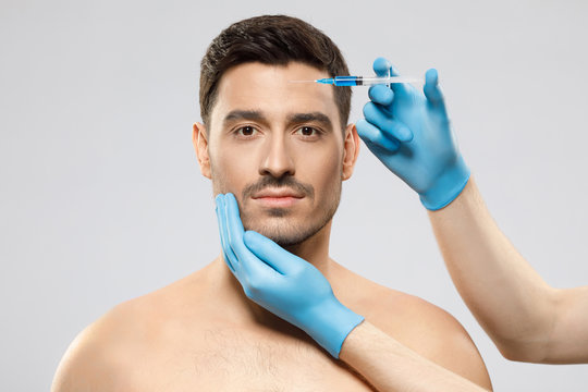 Young man isolated on gray background looking at camera while receiving botox into forehead to prevent sking aging by doctor in blue medical gloves. Face lifting male
