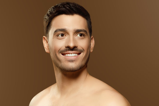 Young handsome man looking aside with smile, standing in front of camera with clear skin, concept of male beauty, isolated on brown background
