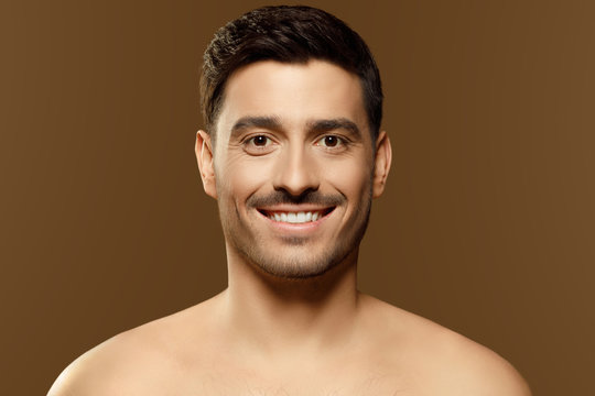 Close-up of beautiful man face looking straight at camera with happy smile, not wearing shirt, concept of skin care, isolated on brown background