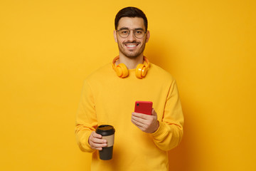 Young smiling guy wearing bright sweater, wireless headphones, holding takeaway coffee cup in one hand and phone in another, isolated on yellow background