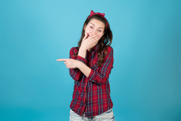 A country girl in a red plaid shirt laughs, covering her mouth and points a finger to the side. Isolated on blue background.