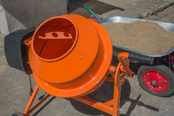 Cement mixer machine is at construction site with wheelbarrow, tools, sand and cement bag. Orange concrete mixer at building construction works.