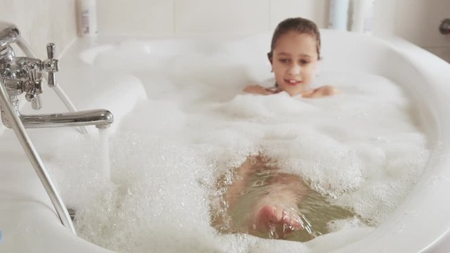 Close-up Of A Pretty Little Girl Squishes Feet In A Bathtub Full Of Foam. Hygiene And Baby Care Concept, HD Still Video Camera, 4x Slow Motion.