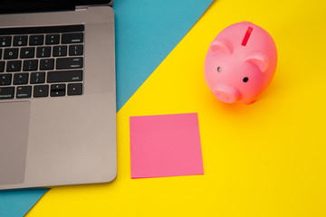Moneybox near laptop and pink sticky note, place for text. Finance and budget concept. Piggy bank in blue color with gadgets and stationery on colorful background.