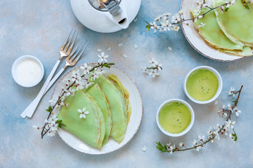 Delicious homemade green pancakes with blossoming cherry tree branches with green matcha tea on sky blue background. Top view, close up