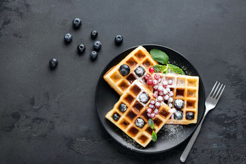 Square belgian waffles with berries and icing sugar on black plate, black background with copy space. Table top view sugar dessert on plate