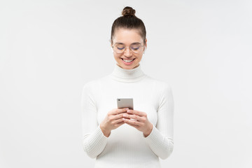 Fototapeta premium Young woman in glasses, holding smartphone in hands, exchanging messages with friends with happy smile, isolated on gray background