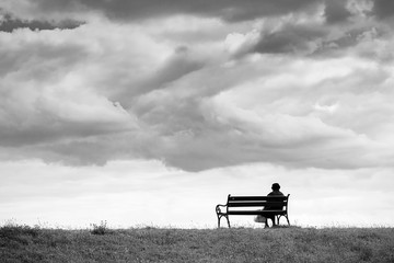 Naklejka premium One woman sits on a bench and looks at the sea. Line horizon and earth on the background of beautiful clouds. Horizontal frame. Black and white image. High contrast.