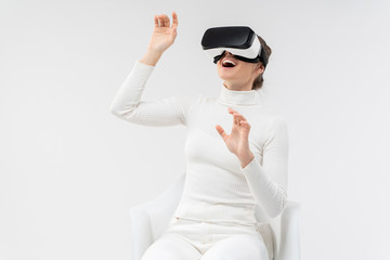 Young woman excited by virtual reality gaming in VR headset on, moving hands in air while sitting in armchair, as if touching objects, isoalted on white background