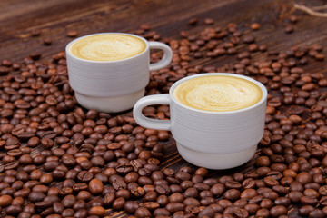 Two white cups of cappuccino on coffee beans heap. Scattered roasted coffee beans on wooden table.
