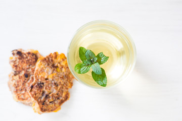 Transparent glass of light yellow green tea with peppermint leaves on white wooden background and two fried pancakes on left out of focus. Biological products breakfast set for healthy life