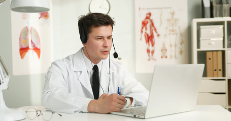 Portrait of a handsome young doctor with headset in front of her laptop, talking with a patient. Medic having videochat and consultation online.