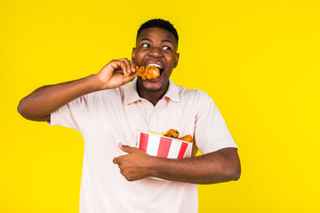 African American young guy eats fast food, chicken leg and a whole bucket in his hands. Emotions of joy and happiness, surprise. Yellow background