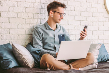 Young businessman in glasses hold, using mobile phone, working on laptop at home office. Stylish hipster man student text message on smart phone. Online communication, remote, distance job on couch