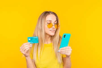 Portrait of a young blonde girl in sunglasses showing plastic credit card while using mobile phone.