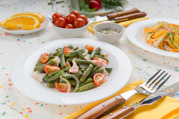 Delicious warm salad with green beans bacon and tomatoes closeup in white plate, healthy vegetable snacks, cereals and orange are served on table