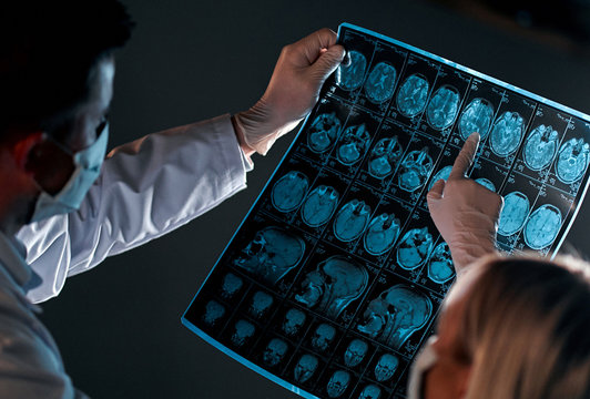 Two doctors in protective masks look and discuss an X-ray or MRI scan of the patient’s brain, isolated on a dark gray background. - Powered by Adobe