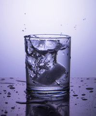 Splash of water in a glass with ice on a gray lilac background. Throw ice while making a cocktail.