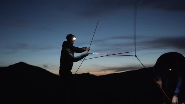 Silhouette of two hikers with head lamps setting up tent in the dark, 4K