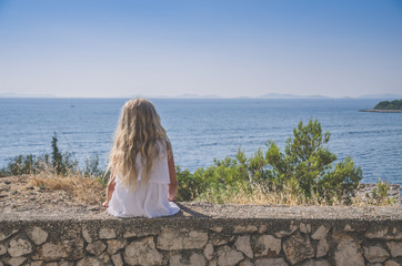 girl with long blond hair sitting alone back view by the sea