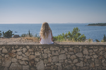 sad girl with long blond hair sitting alone back view by the sea and waiting