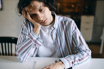 Portrait of sleepy male student in casual clothing sitting at white desk holding hand on his face, having bored look, being tired of doing homework, needs some sleep. People and lifestyle concept