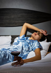 A distraught woman wearing satin pajamas sits in bed with a glass of wine