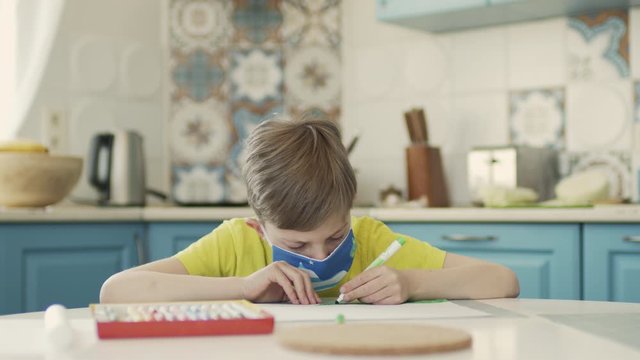 Boy In Medical Mask Sits At Table In The Kitchen And Draws.