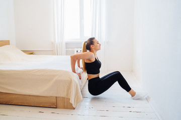 Beautiful athletic girl, wearing black clothes, pumping press indoors at home in a bright light coloured bedroom on a white bed. Isolated at home. Fitness without gym. Healhty sportive concept