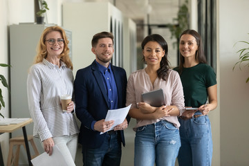 Portrait of happy smiling diverse mixed race employees team holding equipment standing in office, holding cup of coffee and tablet, posing for company photo together, looking at camera.