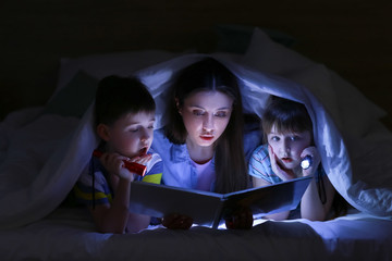 Woman and her little children reading book under blanket at night
