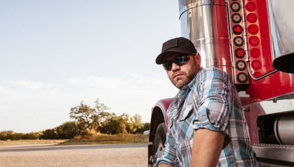 Casual dressed trucker with plaid shirt and baseball cap sits next to red big rig. Commercial...