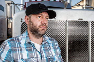 Worried semi truck driver wearing plaid shirt and black baseball hat sits in front of big rig....