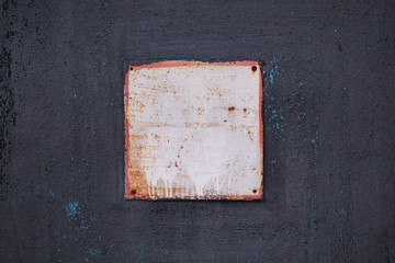 A square aluminum sign with a red frame on an old painted metal black wall. The sign is rusted and painted white. The paint on the black wall is cracked and peeling. Industrial background, backdrop.