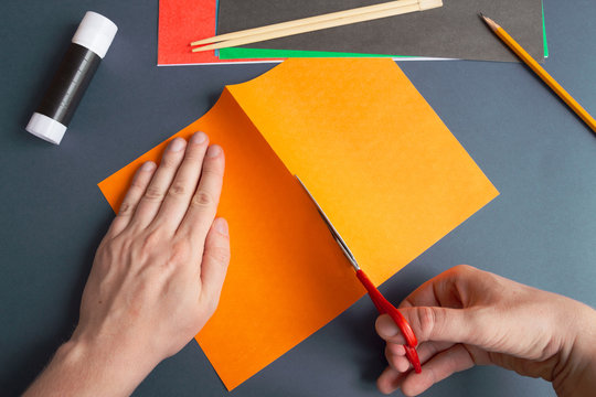 Paper Halloween Jack O'Lantern Step-by-step Instruction. Step 3: Person Cuts An Orange Sheet Of Paper Into Two Parts With Scissors. DIY Halloween Decoration Theme.