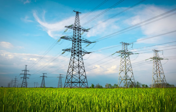 High voltage lines and power pylons in a flat and green agricultural landscape on a sunny day with clouds in the blue sky. Cloudy and rainy. Wheat is growing - Powered by Adobe