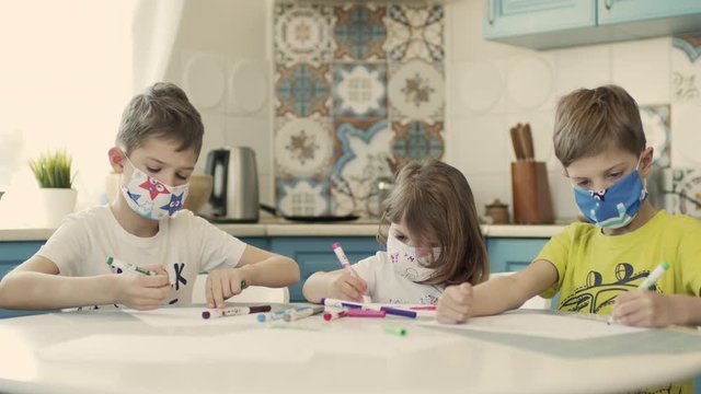 Two Boys And Their Little Sister In Multicolored Medical Masks Draw At The Table In The Kitchen.